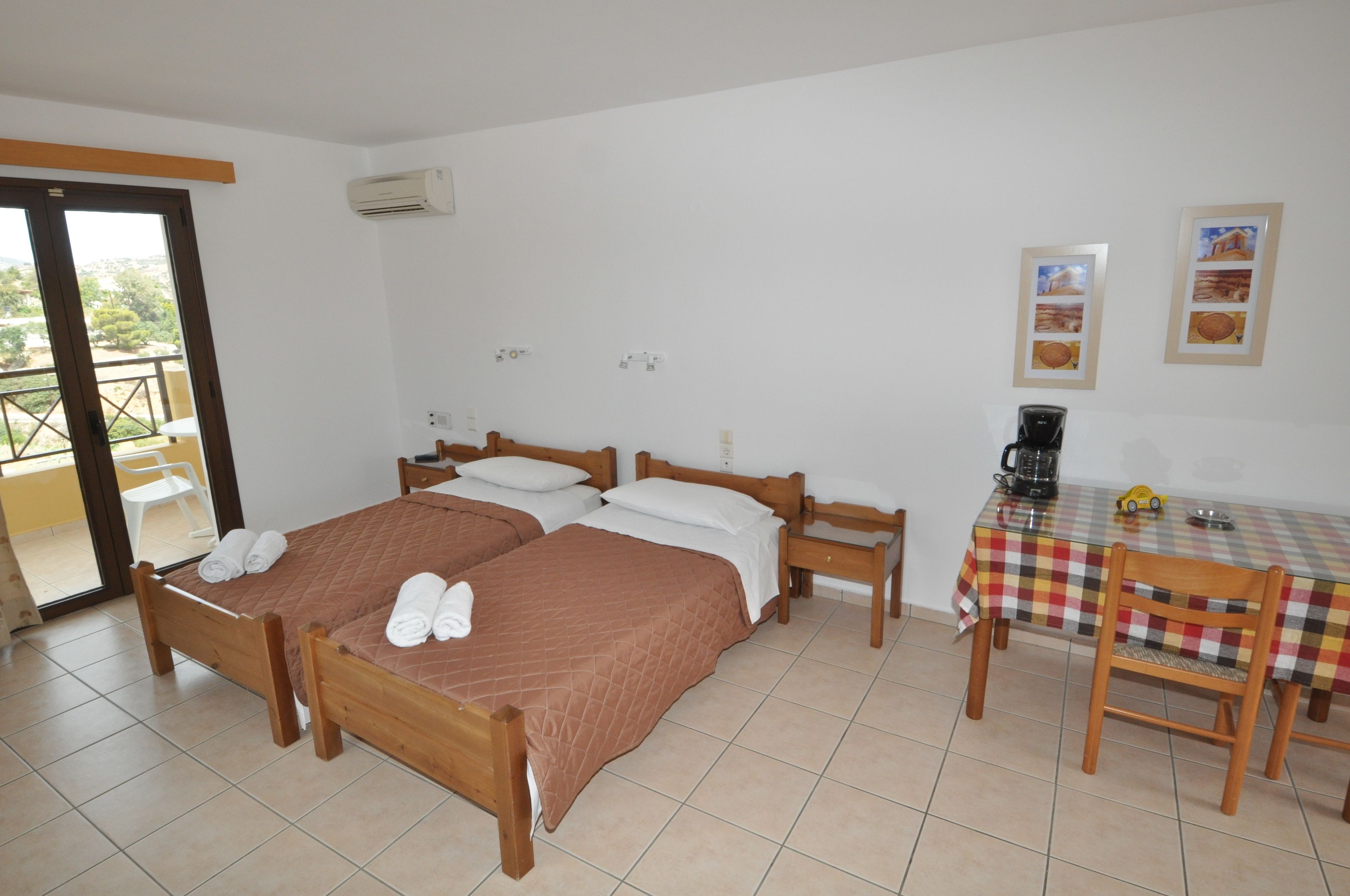 A room with two wooden beds, a dining table with a checkered tablecloth, a chair, and a coffee maker.