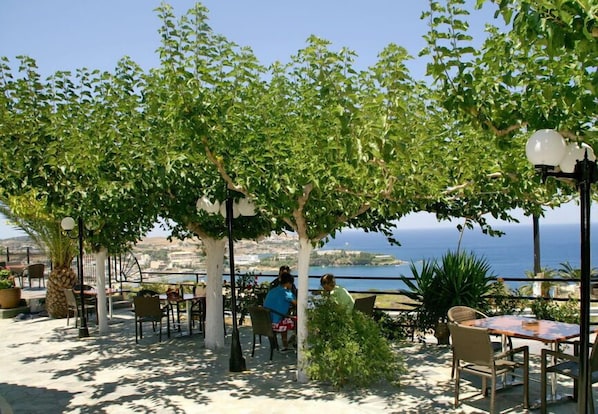 An outdoor seating area with tables and chairs, surrounded by trees and overlooking a scenic view of the sea.
