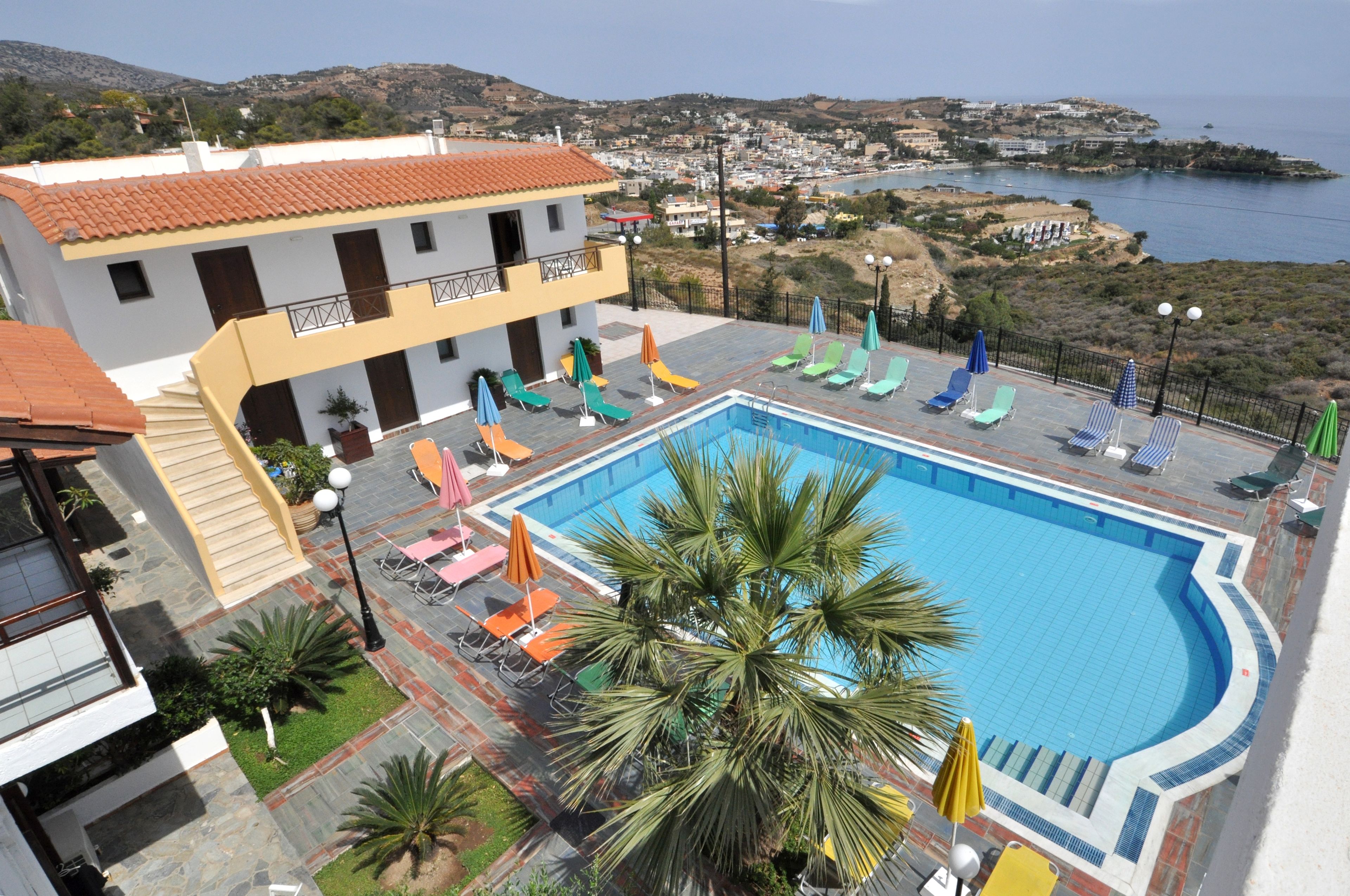 A swimming pool with lounge chairs and umbrellas, surrounded by a building with a tiled roof and a view of the sea and hills.