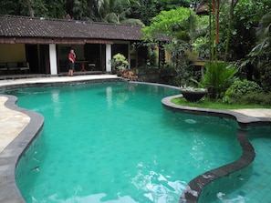 A swimming pool with a curved edge, surrounded by lush greenery and a building with a tiled roof. - Villa Jineng Ubud Bali (Ubud)