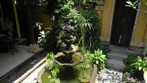 A multi-tiered stone fountain surrounded by various green plants, set in a courtyard with a tiled floor and a stone wall in the background. - Villa Jineng Ubud Bali (Ubud)
