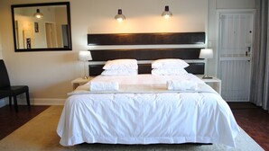 Una cama bien hecha con sábanas blancas, dos lámparas de noche, un espejo grande y una puerta con marco blanco.