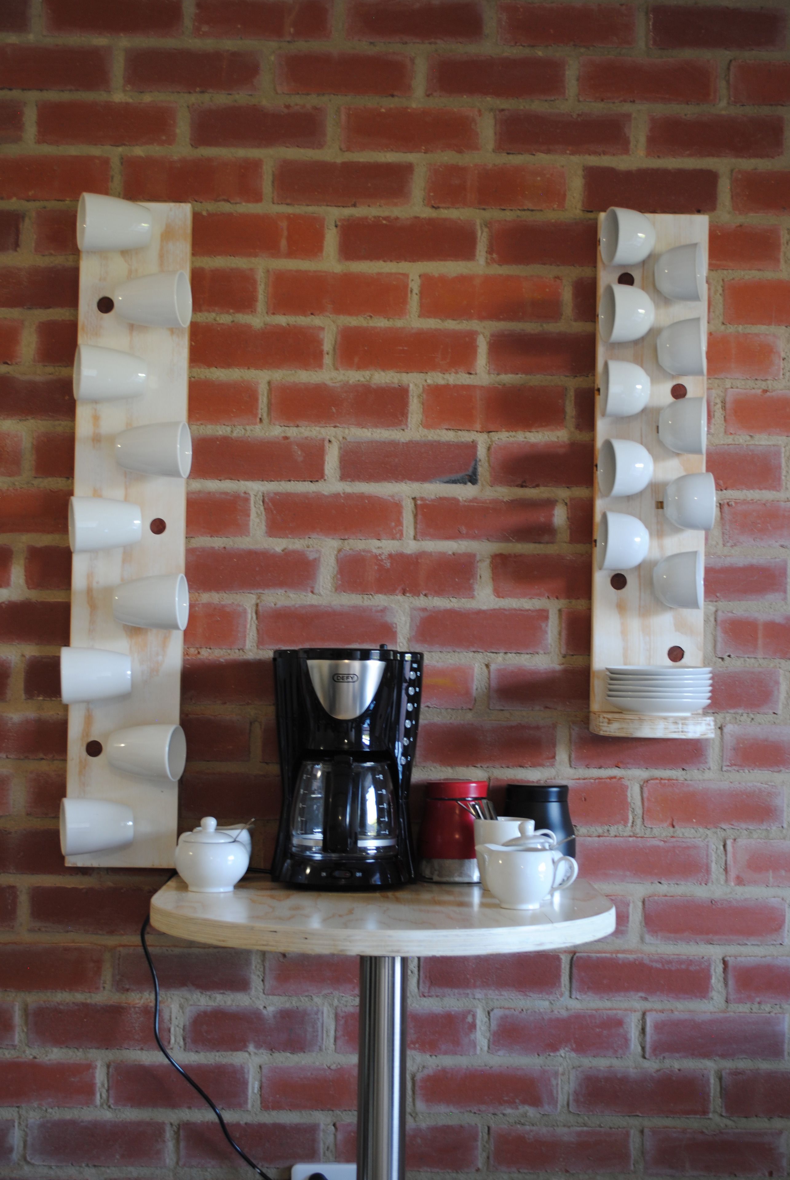 Una estación de café con una cafetera, tazas y una pequeña mesa frente a una pared de ladrillo.