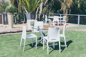 An outdoor patio area with white chairs and tables, surrounded by a metal fence and greenery.