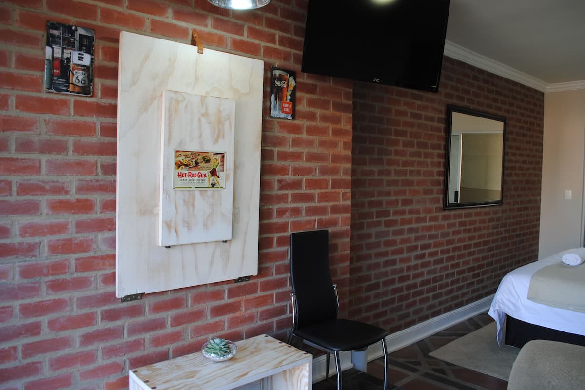 A hotel room with a brick wall, a bed, a black chair, a small table, and a mounted TV.