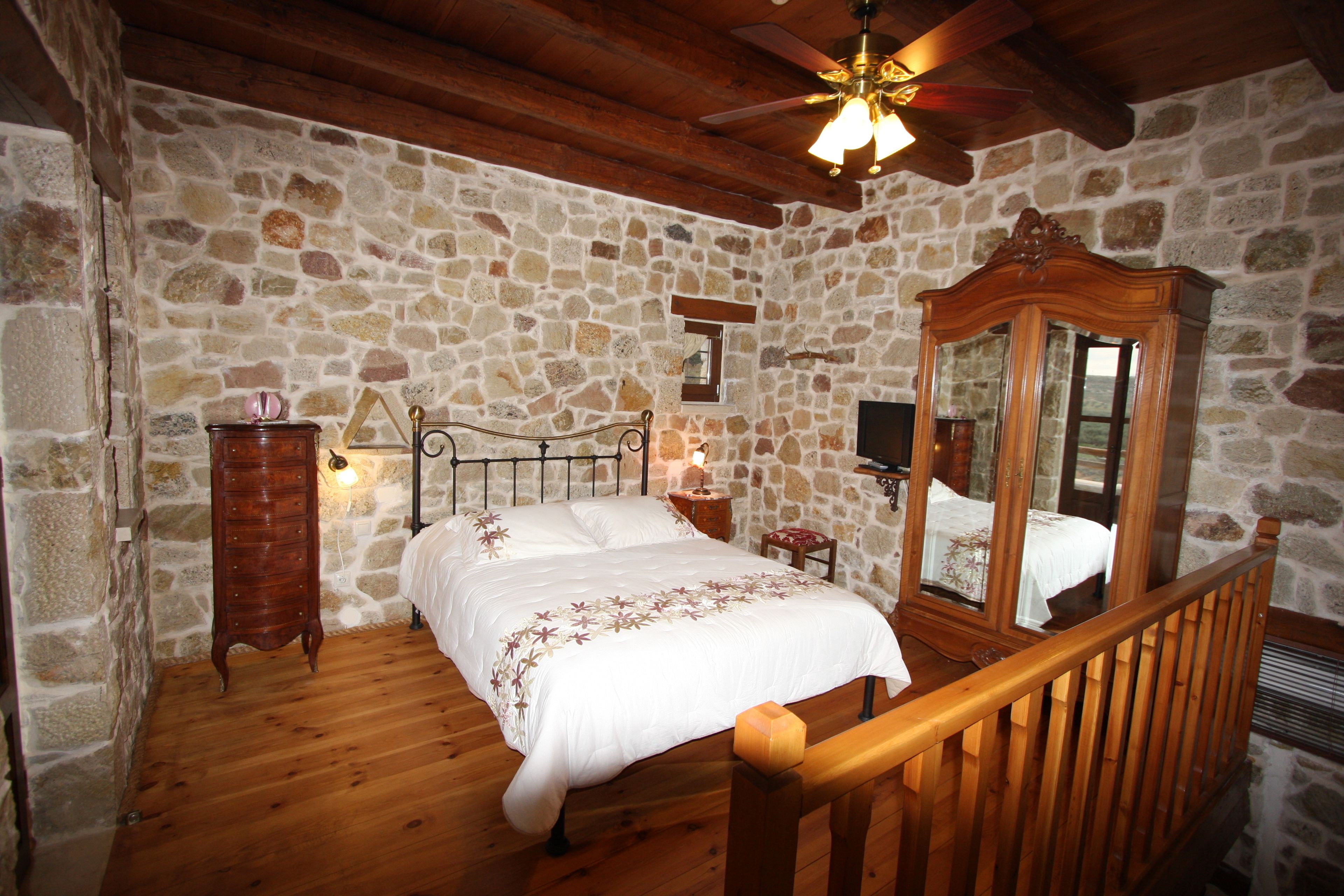 Ein Schlafzimmer mit Steinwand, Holzboden, einem Bett mit Holz-Kopfteil, einer Kommode aus Holz, einem Spiegel und einem Deckenventilator.
