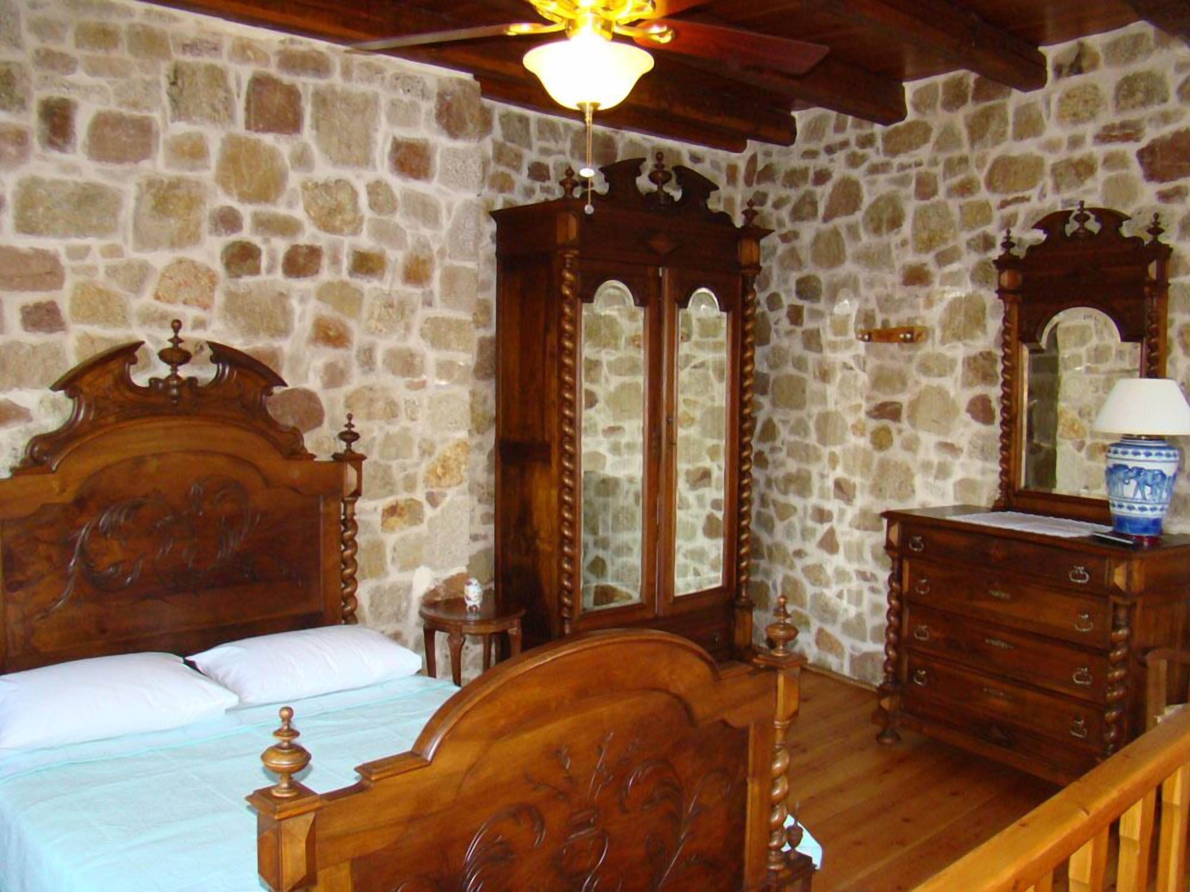 A bedroom with a stone wall, a wooden bed with a headboard, a dresser, a mirror, and a ceiling fan.
