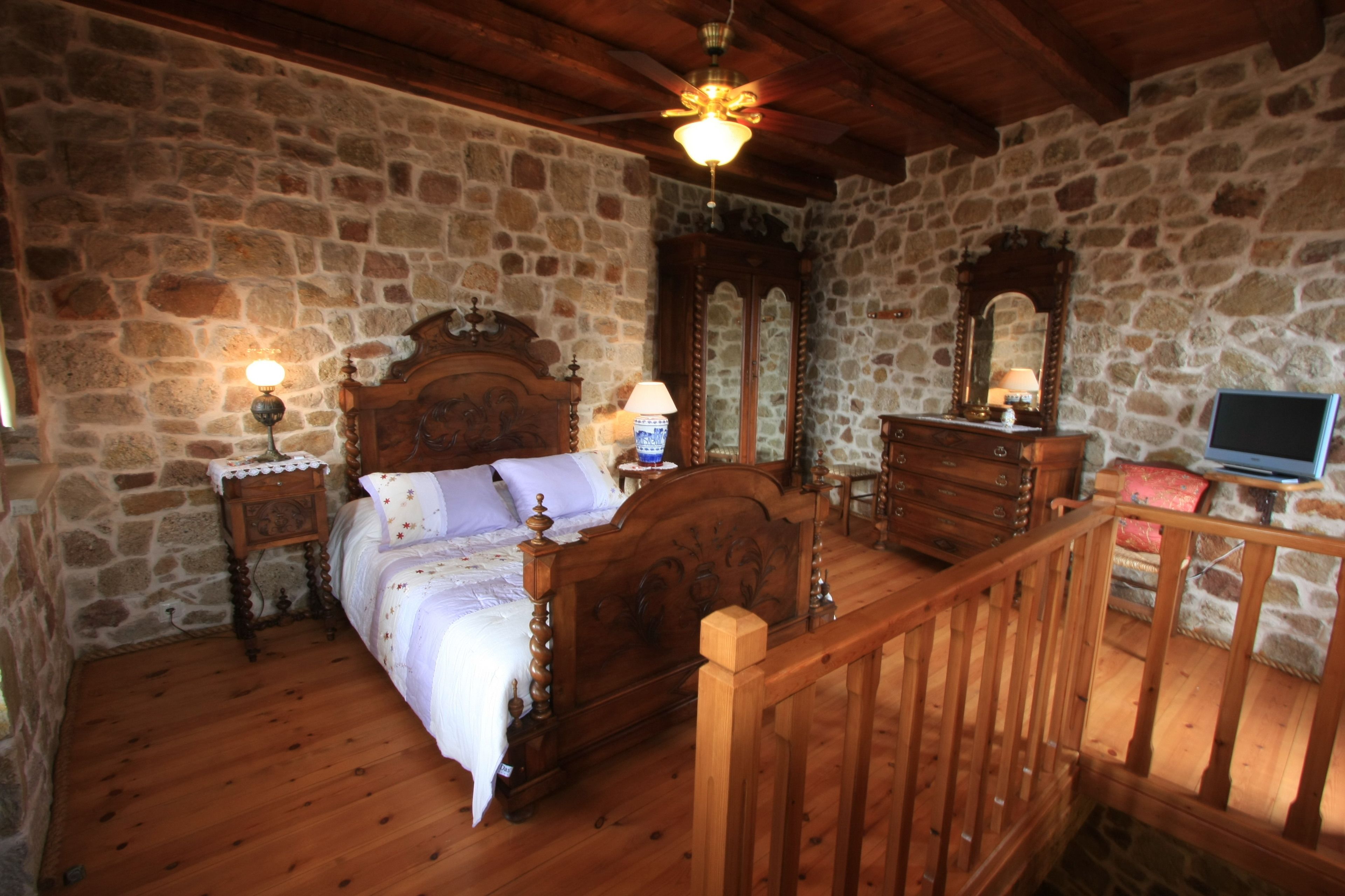 A bedroom with stone walls, a wooden bed, a nightstand with a lamp, a dresser, and a television.