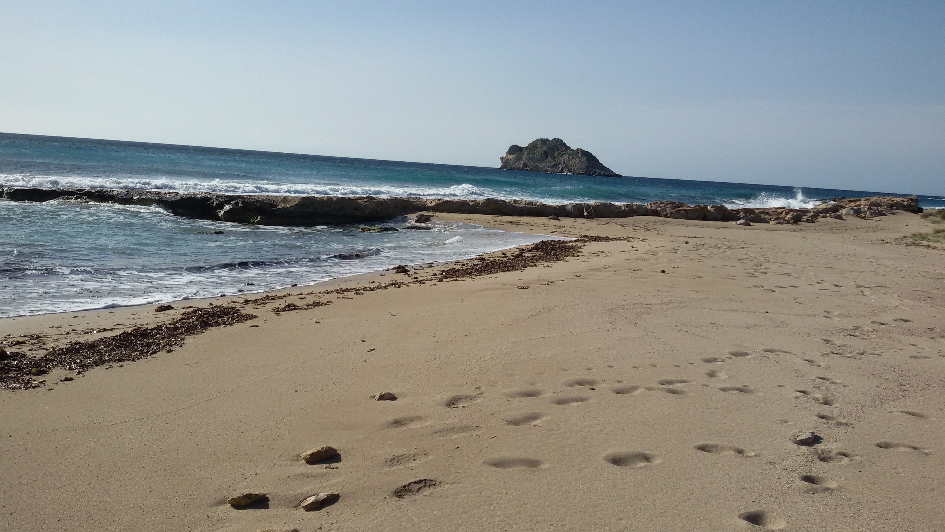 A sandy beach with footprints and seaweed, leading to rocky cliffs and a clear sky.