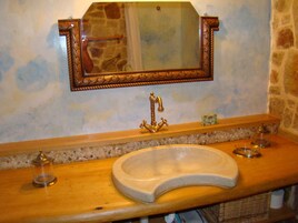 Un baño con pared de piedra, tocador de madera, espejo grande, grifo dorado y jabonera.