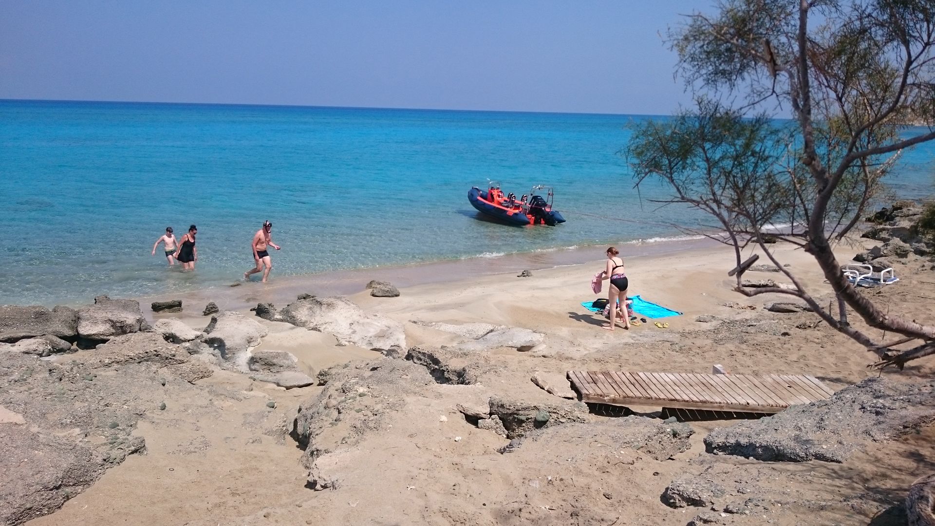 A beach with clear blue water, rocky foreground, and a boat in the distance.