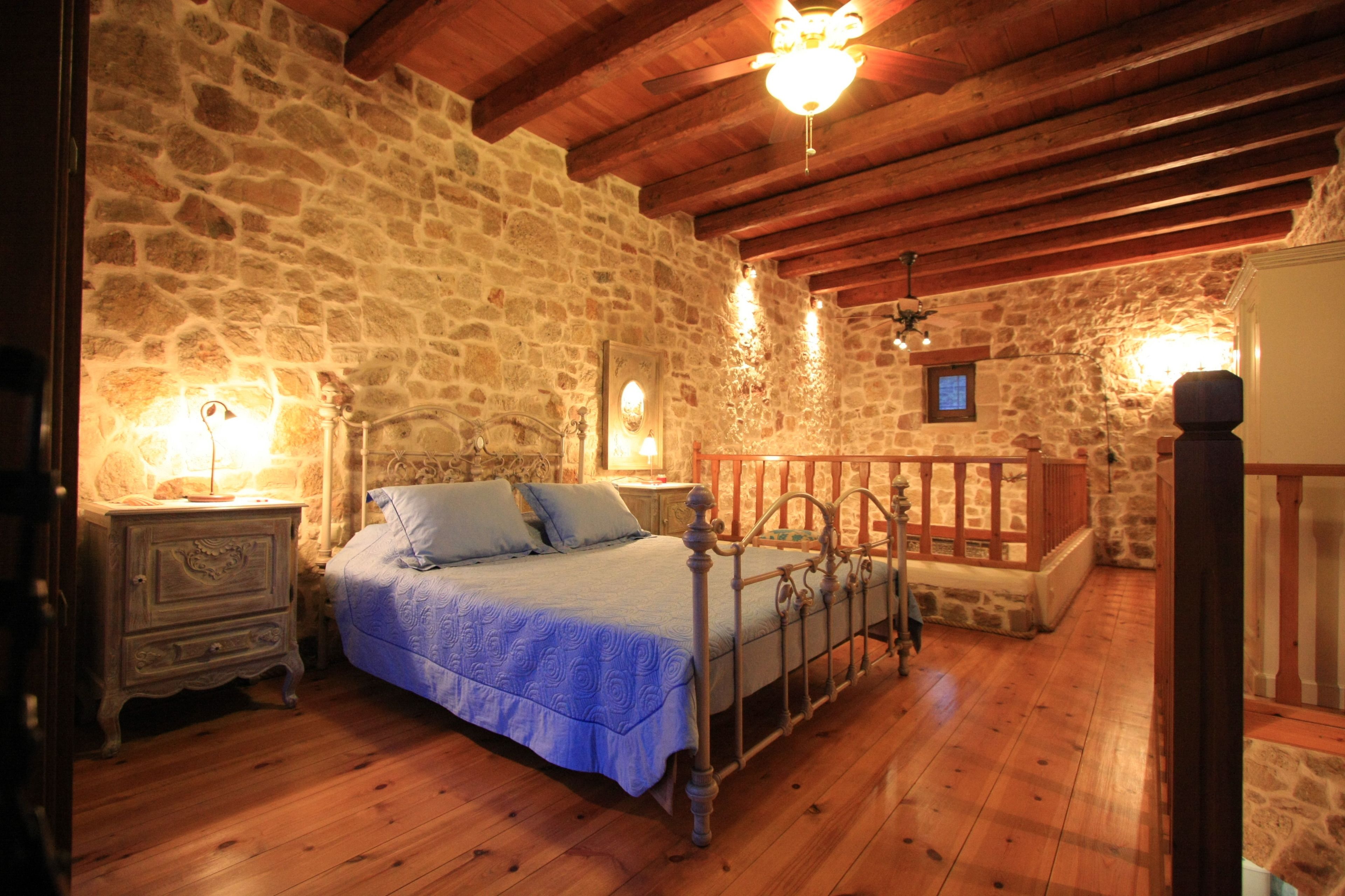 Une chambre avec un mur en pierre, un plancher en bois, un lit avec un cadre en métal, une table de chevet et une lampe suspendue.