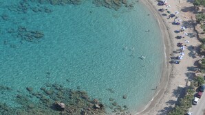 Luchtbeeld van een strand met helder turquoise water, een rotsachtige kustlijn en een rij strandparasols langs de oever.