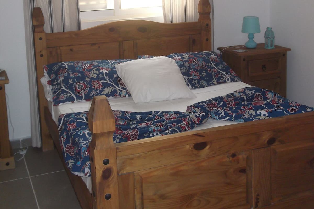 Una cama de madera con ropa de cama floral, una mesita de noche con una lámpara y una ventana con cortinas.