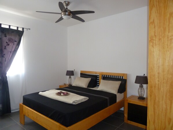 A bedroom with a wooden bed, two bedside tables with lamps, a ceiling fan, and a window with curtains.