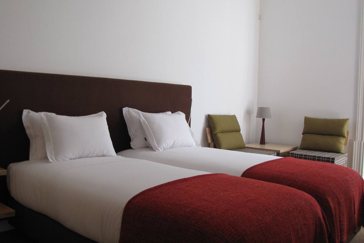 A hotel room with two beds, a nightstand, a lamp, and a chair.