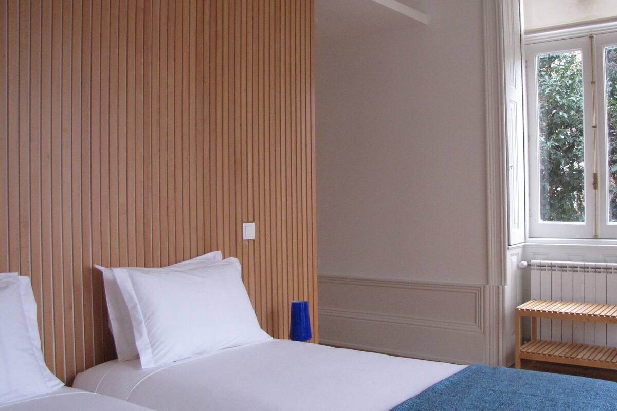 A bedroom with a wooden headboard, a window with a view of greenery, a radiator, and a small wooden stool.