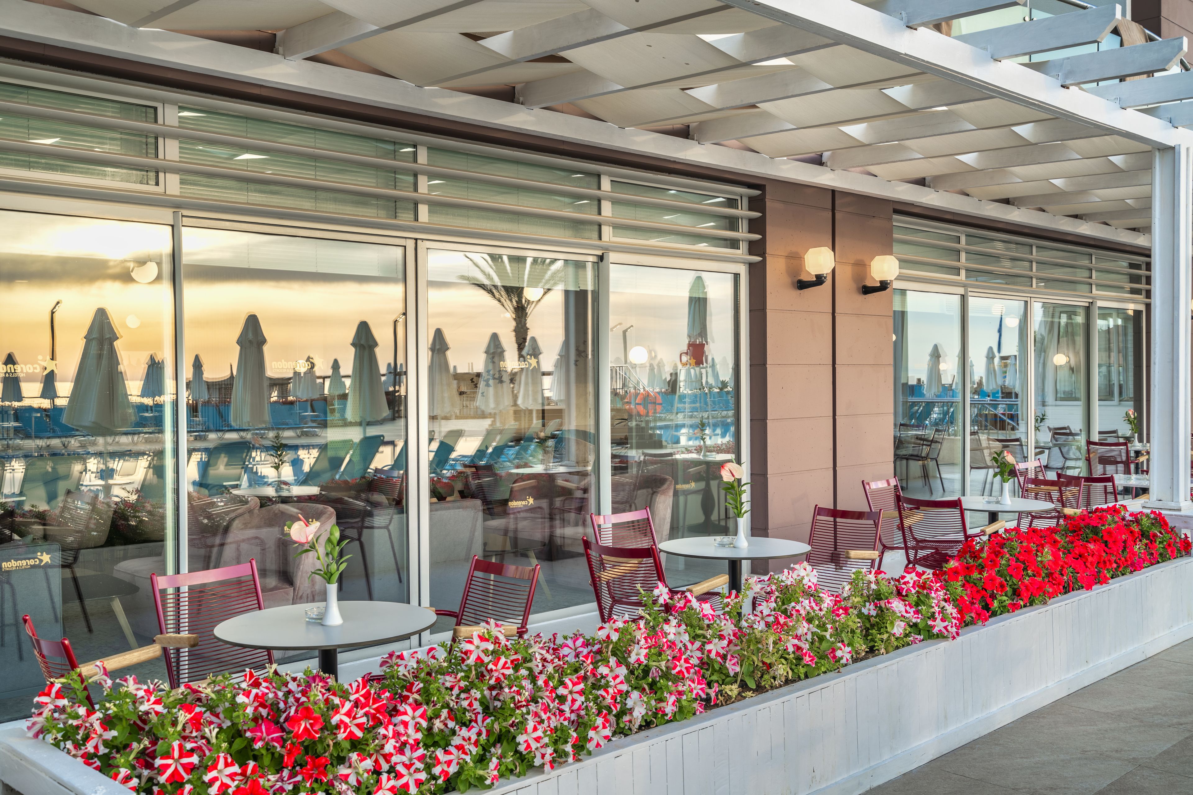 Un área de estar al aire libre con mesas y sillas, amplias ventanas con vistas a la ciudad y un parterre con flores rojas y rosas.