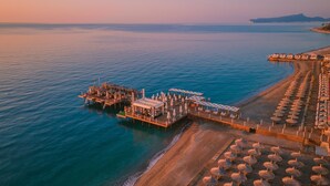 Vista aérea de un complejo turístico costero con un muelle que se adentra en el mar, con varios niveles de asientos al aire libre y sombrillas.
