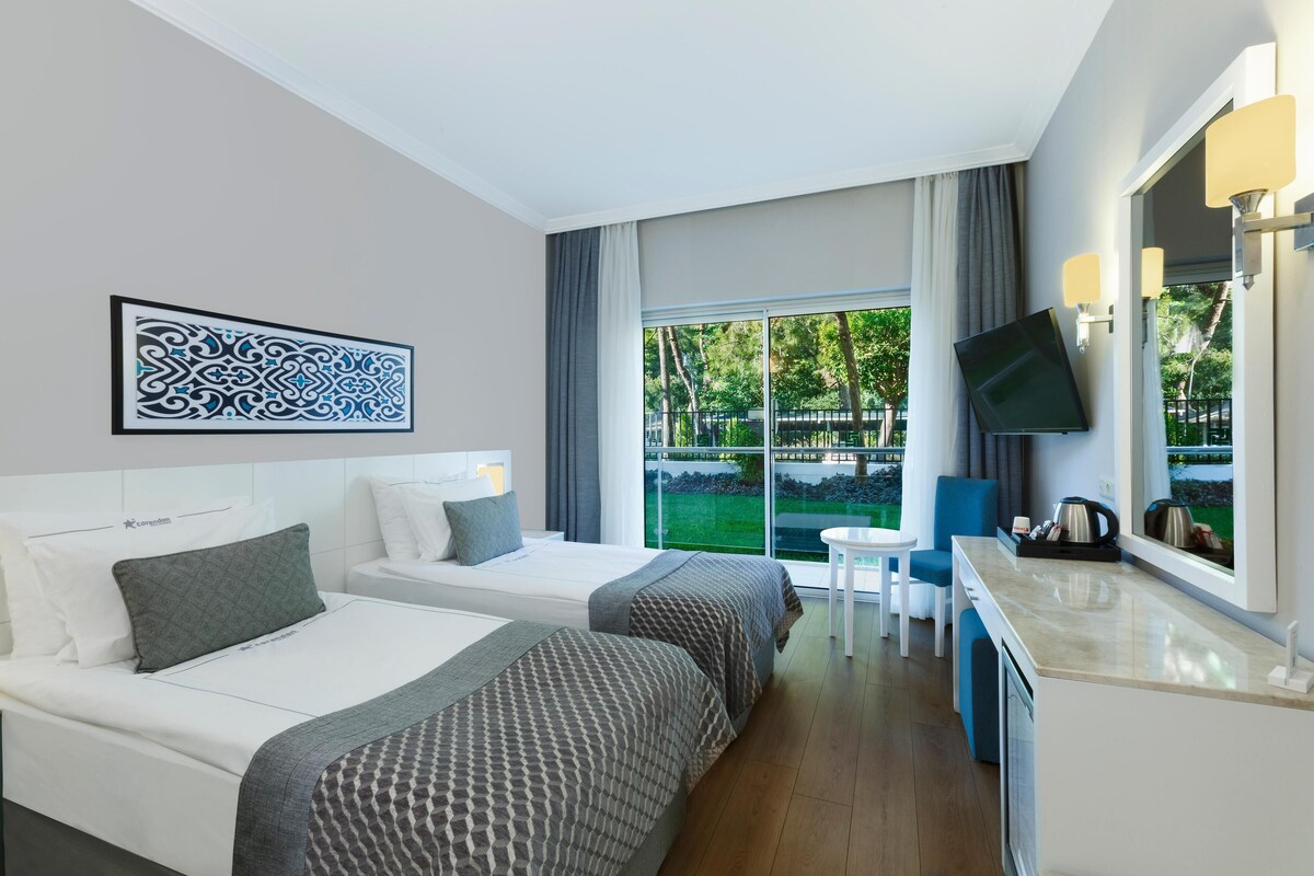 A hotel room with two beds, a TV, a small table, and a view of the outdoors.