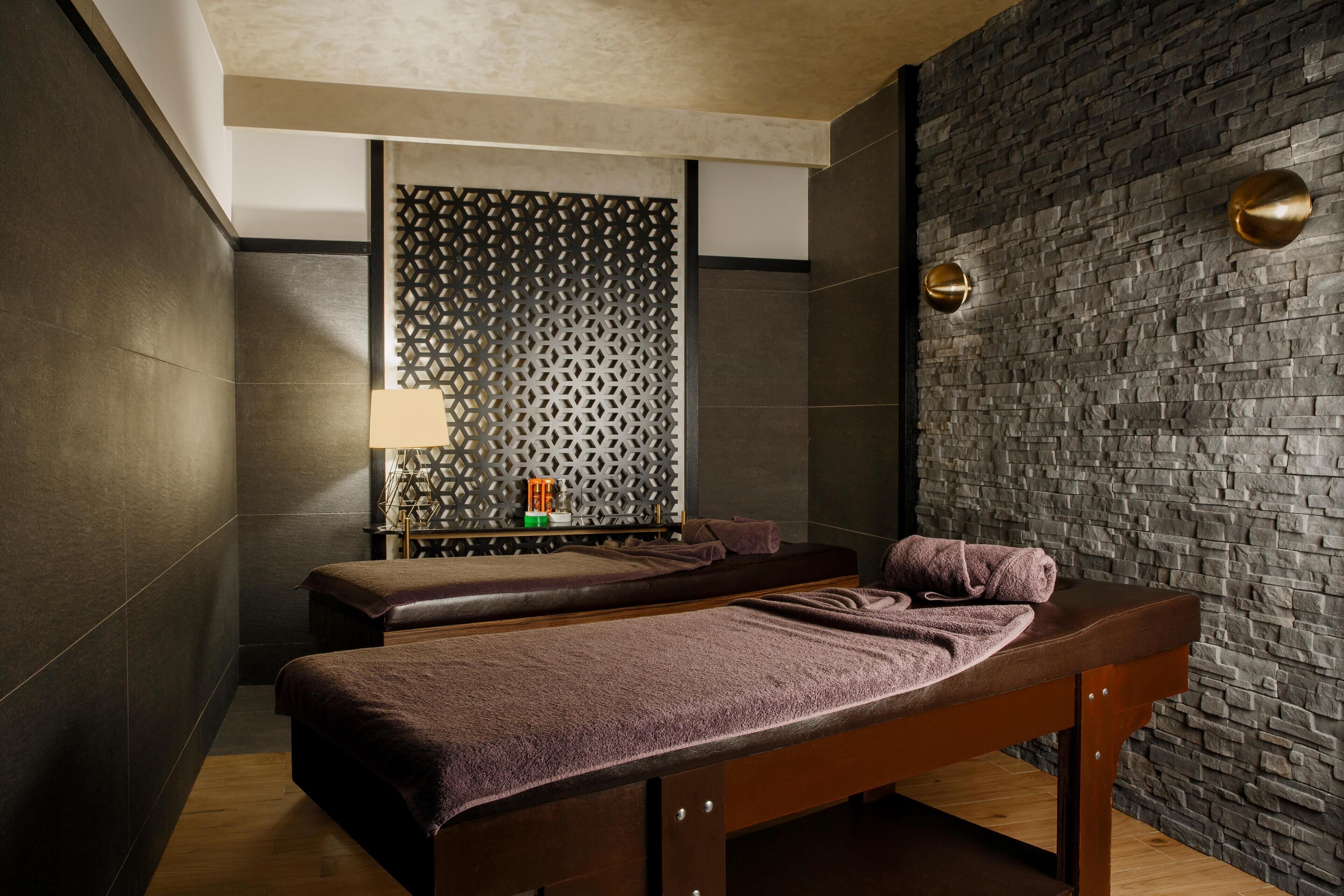 Ein Spa-Raum mit zwei Holzwettenmassagetischen, einer Wand mit Muster und einer kleinen Ablage mit Lampe und Pflegeprodukten.