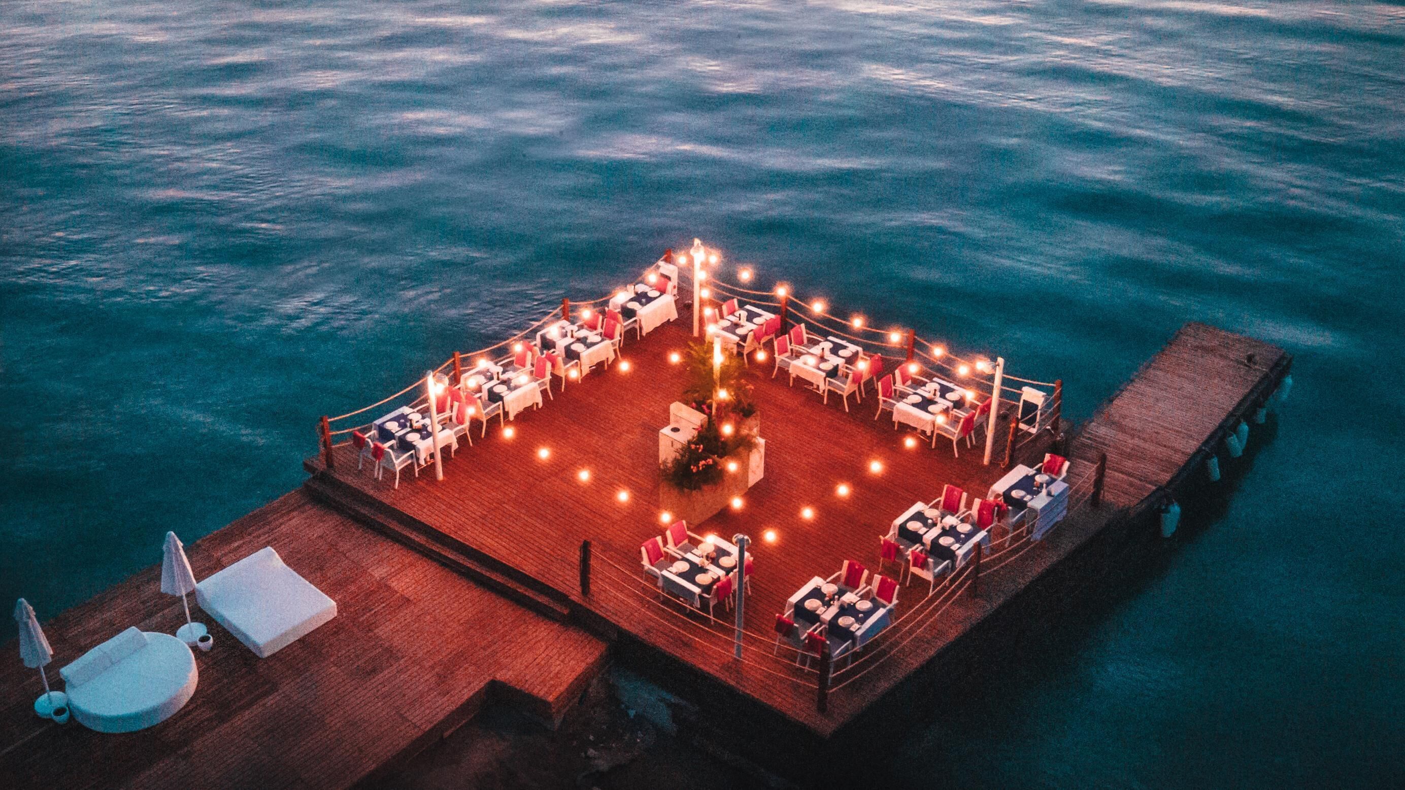 An outdoor dining area on a wooden deck with tables and chairs arranged around a central fire pit.