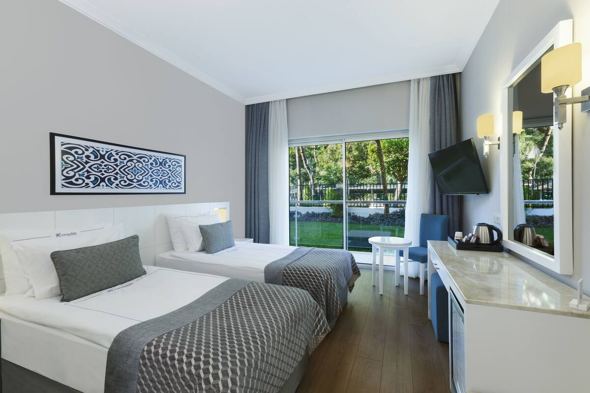 A hotel room with two beds, a TV, a small table, and a window with a view of greenery.