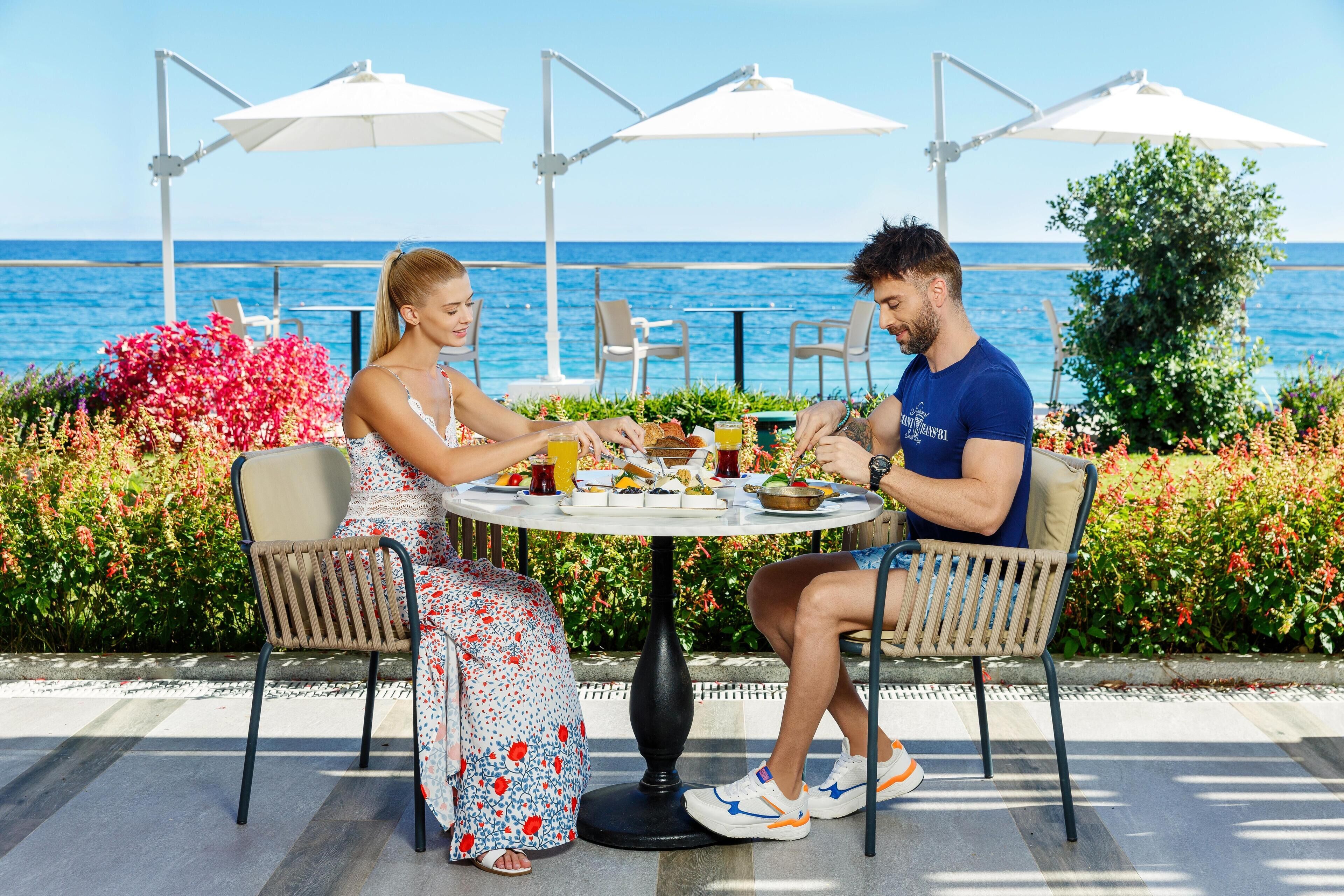 Una pareja cenando en una mesa al aire libre con vista al mar, rodeada de sombrillas y plantas.