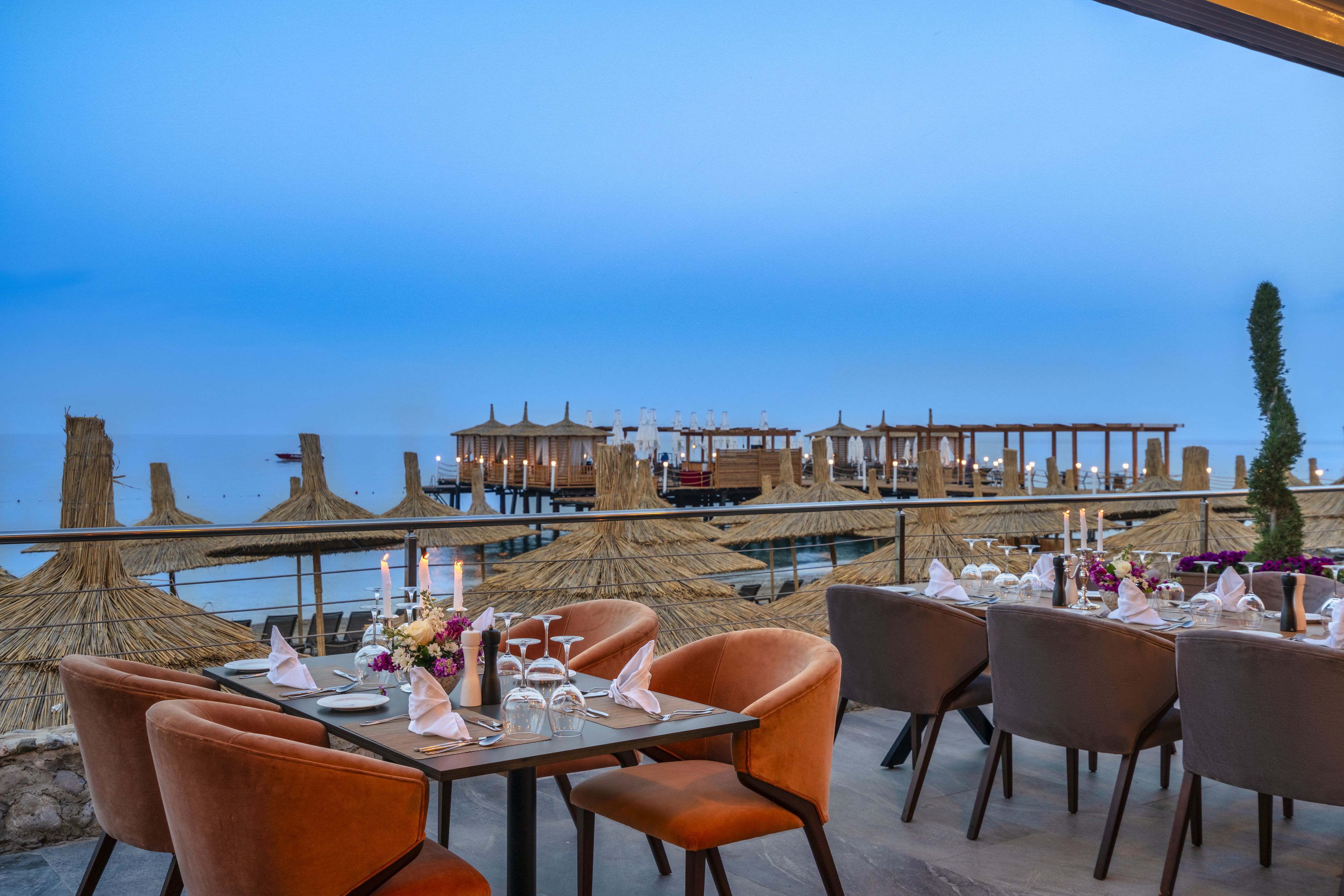 An outdoor dining area with tables set for two, overlooking a waterfront with umbrellas and a pier.