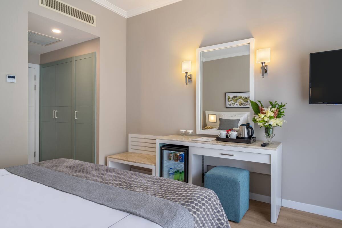 A modern hotel room with a bed, a desk with a mirror, a small fridge, a TV, and a bedside table with a vase of flowers.
