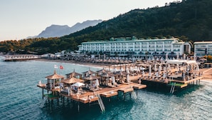 Un hotel frente al mar con varios niveles de balcones con vistas al océano, una zona de playa con sombrillas y montañas al fondo.