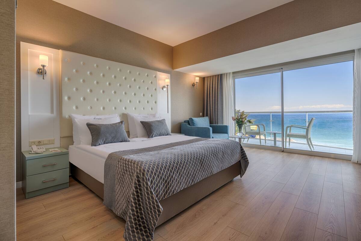 A modern hotel room with a large bed, a nightstand, a seating area with chairs, and a balcony with a view of the ocean.