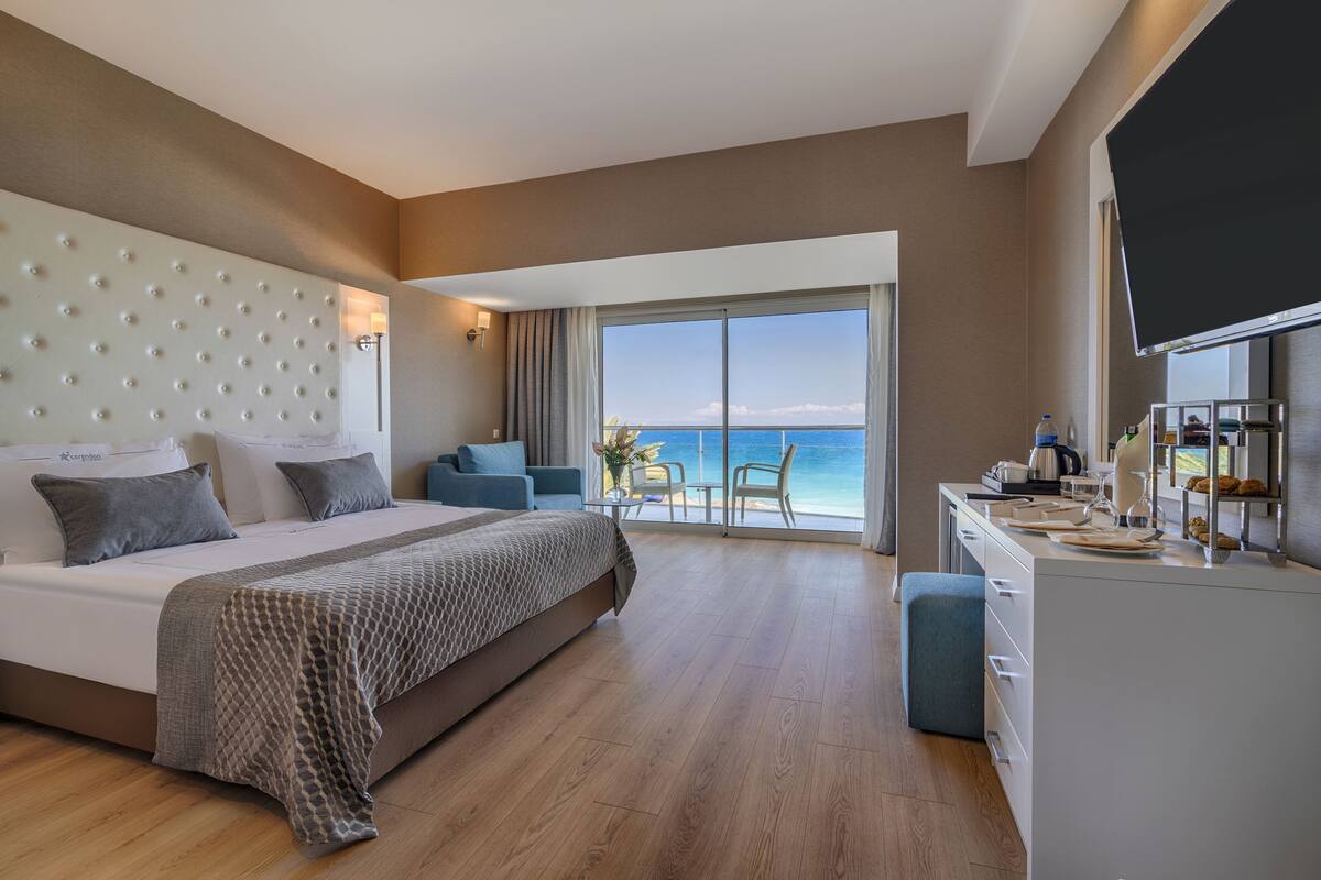 A modern hotel room with a large bed, a desk, and a balcony with a view of the sea.