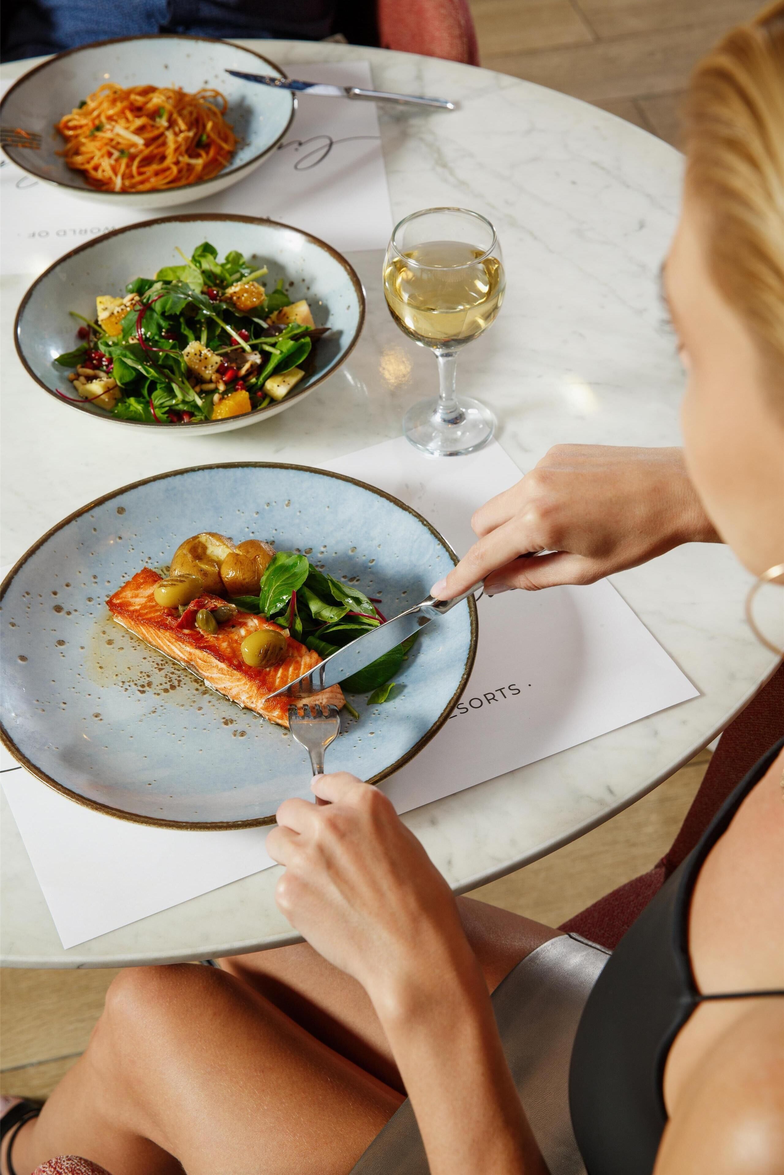 Una mesa de comedor con platos de comida, una copa de vino y una persona cortando una porción de pizza.