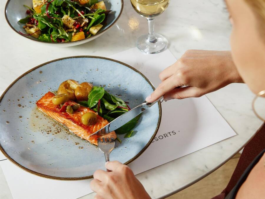 Una mesa de comedor con platos de comida, una copa de vino y una persona cortando una porción de pizza.