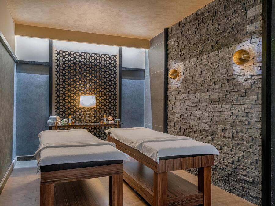 A spa room with two wooden massage tables, a geometric patterned wall, and a stone wall with a light fixture.