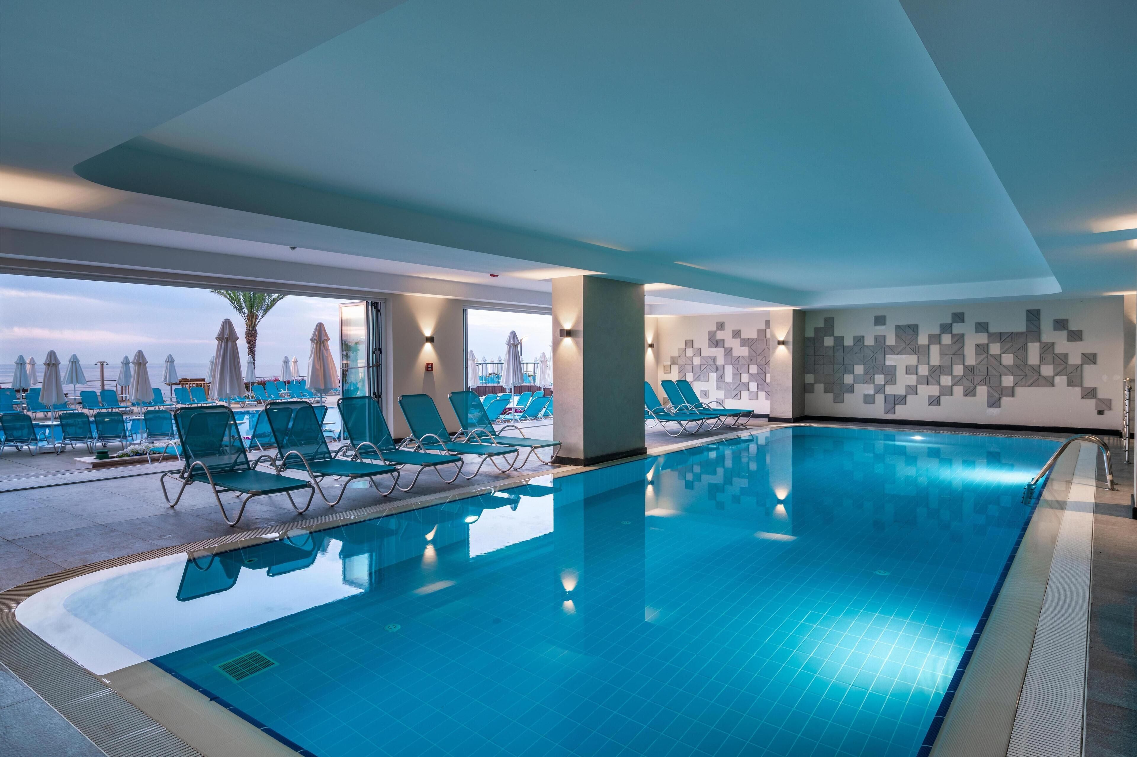 Una piscina interior con azulejos azules, rodeada de sillas de descanso y sombrillas, y vista a la ciudad a través de amplios ventanales.