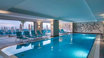 Una piscina interior con azulejos azules, rodeada de sillas de descanso y sombrillas, y vista a la ciudad a través de amplios ventanales.