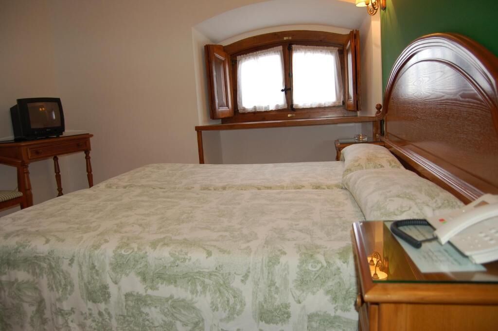 A hotel room with a bed, a bedside table with a telephone, a small table with a television, and a window with curtains.