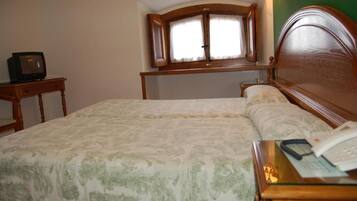 A hotel room with a bed, a bedside table with a telephone, a small table with a television, and a window with curtains.