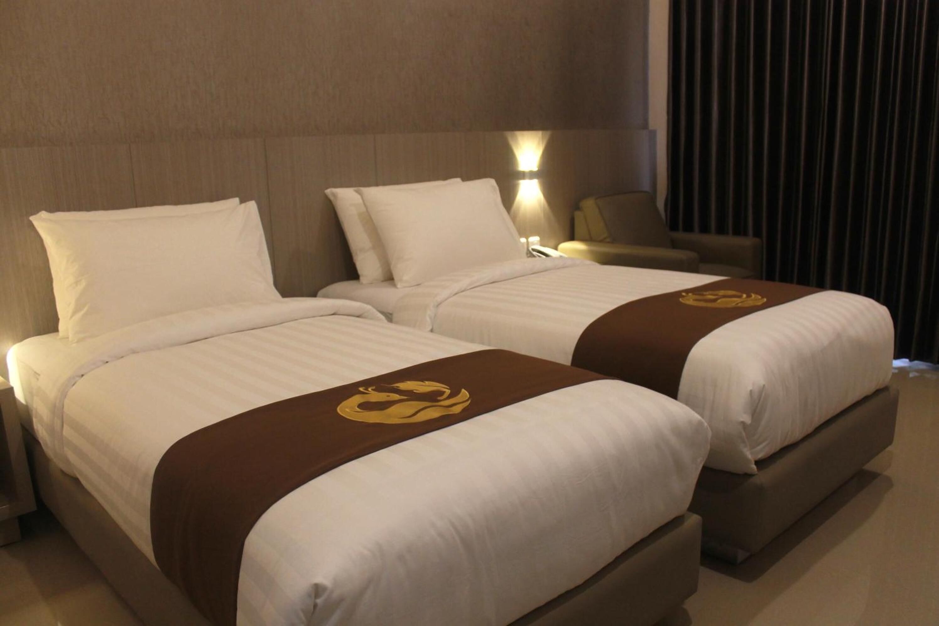 A hotel room with two beds, each with white linens and a brown headboard. There is a nightstand with a lamp and a chair in the corner.