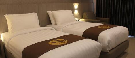 A hotel room with two beds, each with white linens and a brown headboard. There is a nightstand with a lamp and a chair in the corner.