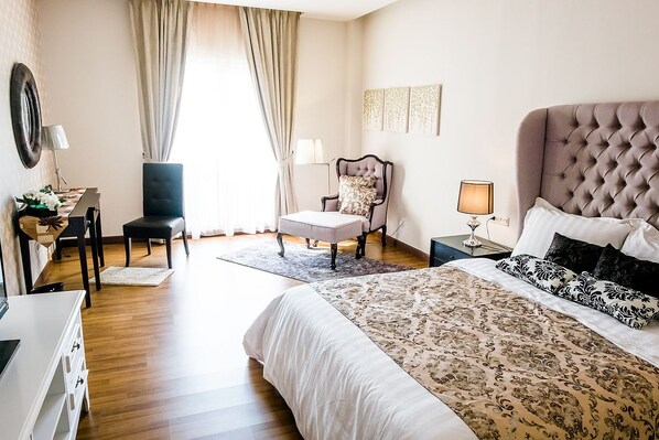 Habitación de hotel con una cama grande, un escritorio con silla, una mesita y un sillón con cojín.