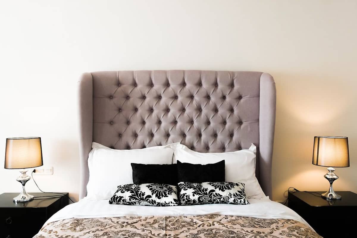 Una cama bien hecha con un cabecero capitonado, dos mesitas de noche con lámparas y una colcha estampada.