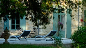 Un bâtiment en pierre avec une porte et des fenêtres vertes, des tables et des chaises en métal à l’extérieur, et un arbre au premier plan.