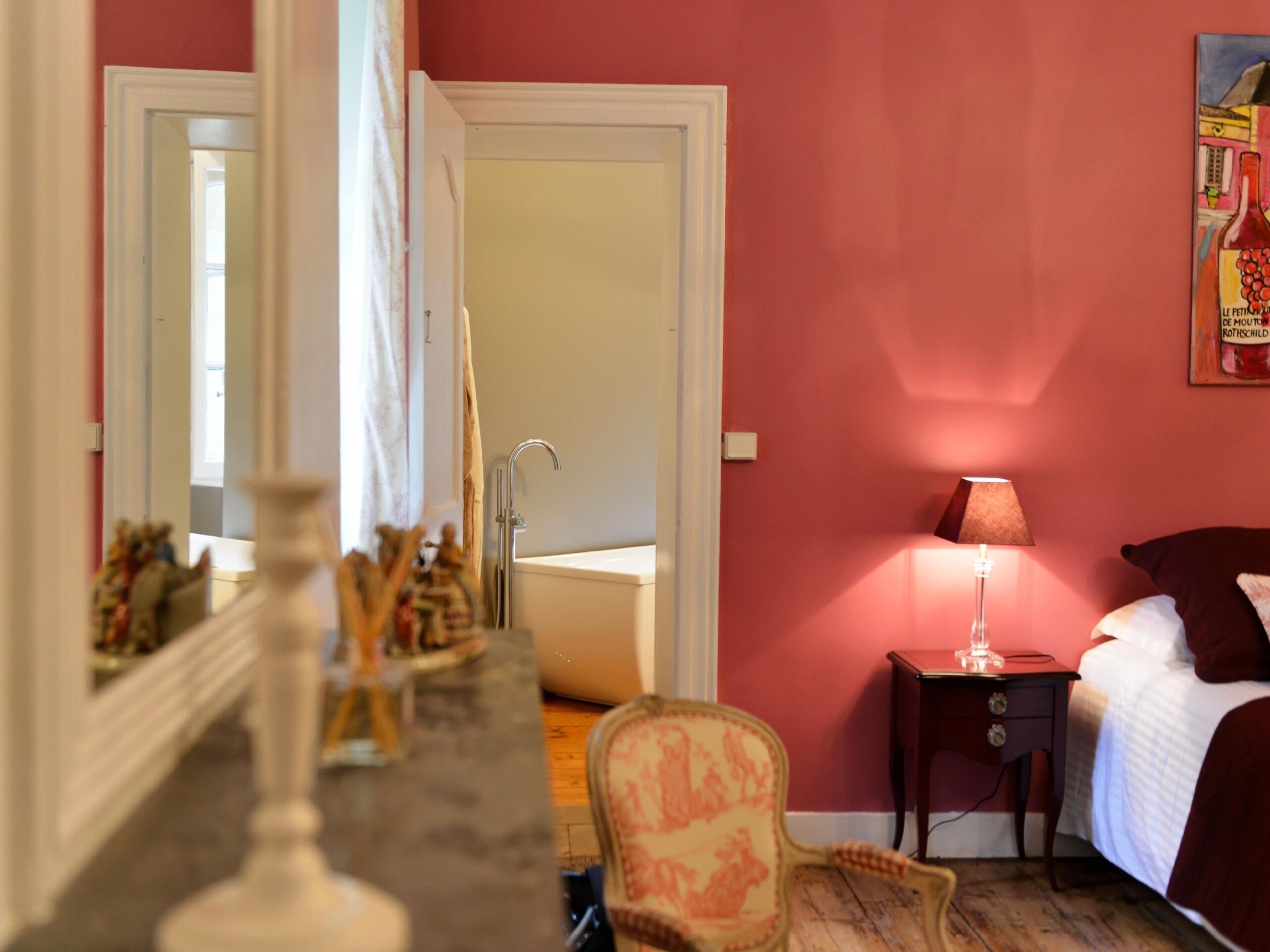 Un dormitorio con cama, mesita de noche, lámpara, sillón y un baño visible a través de una puerta abierta.