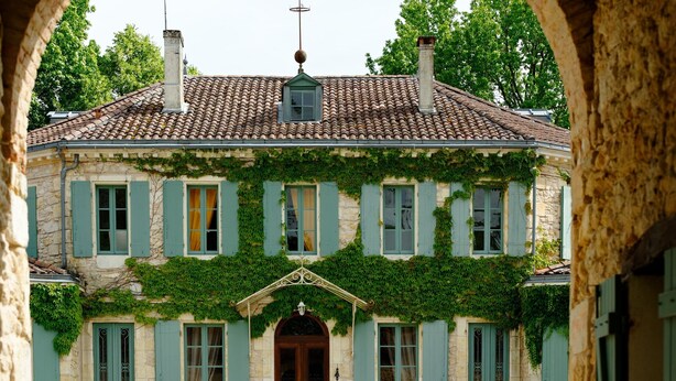 Un bâtiment historique à la façade en pierre, aux volets verts et doté d’un jardin où poussent des plants de lavande.