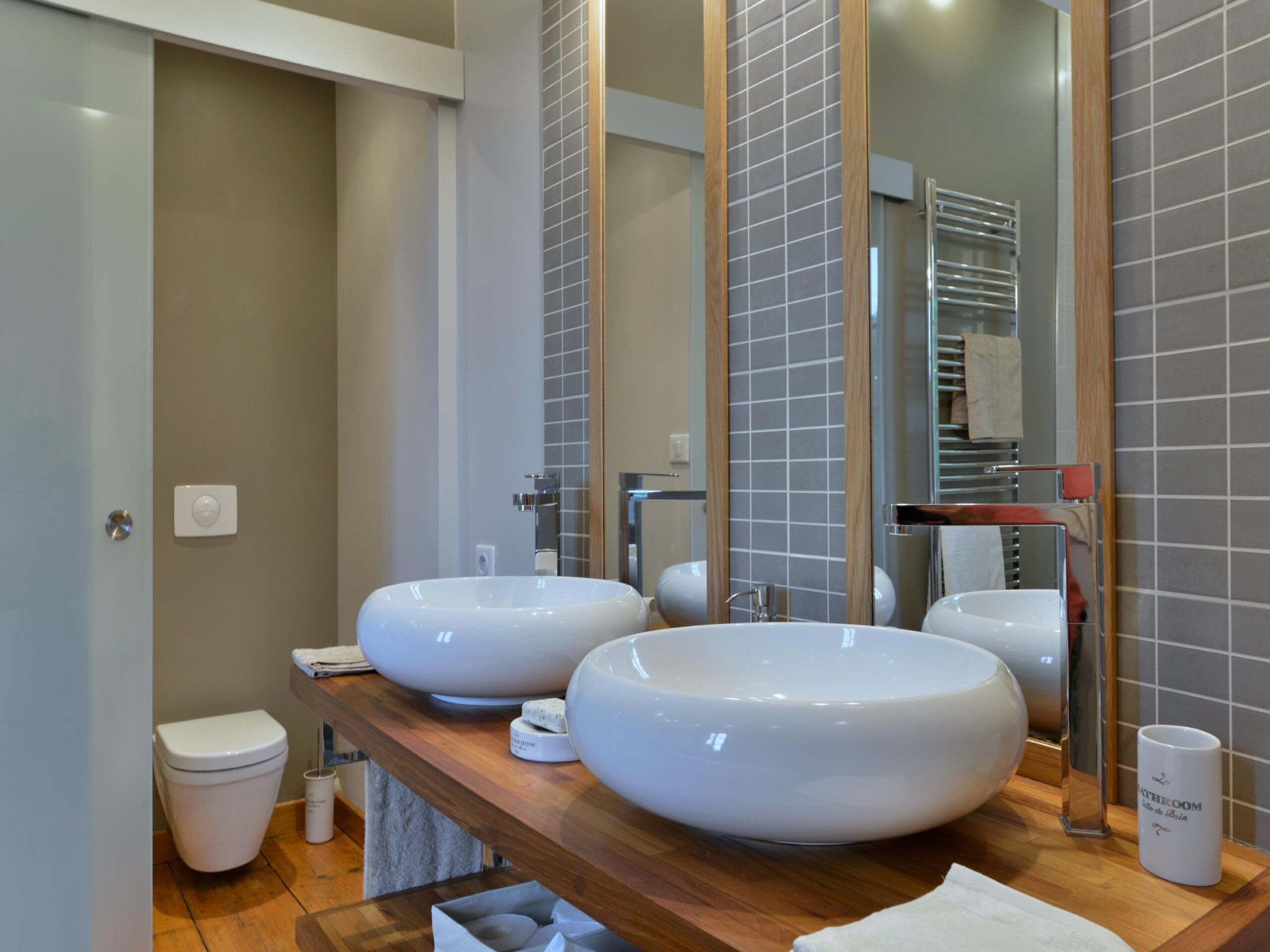 Une salle de bain moderne avec deux grands lavabos blancs, un meuble-lavabo en bois, des toilettes et un miroir avec un cadre en bois.
