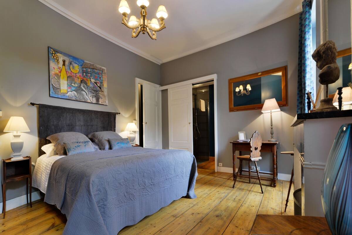Ein Schlafzimmer mit einem großen Bett, einer Kristallleuchtre, einem Gemälde und Holzdielen.