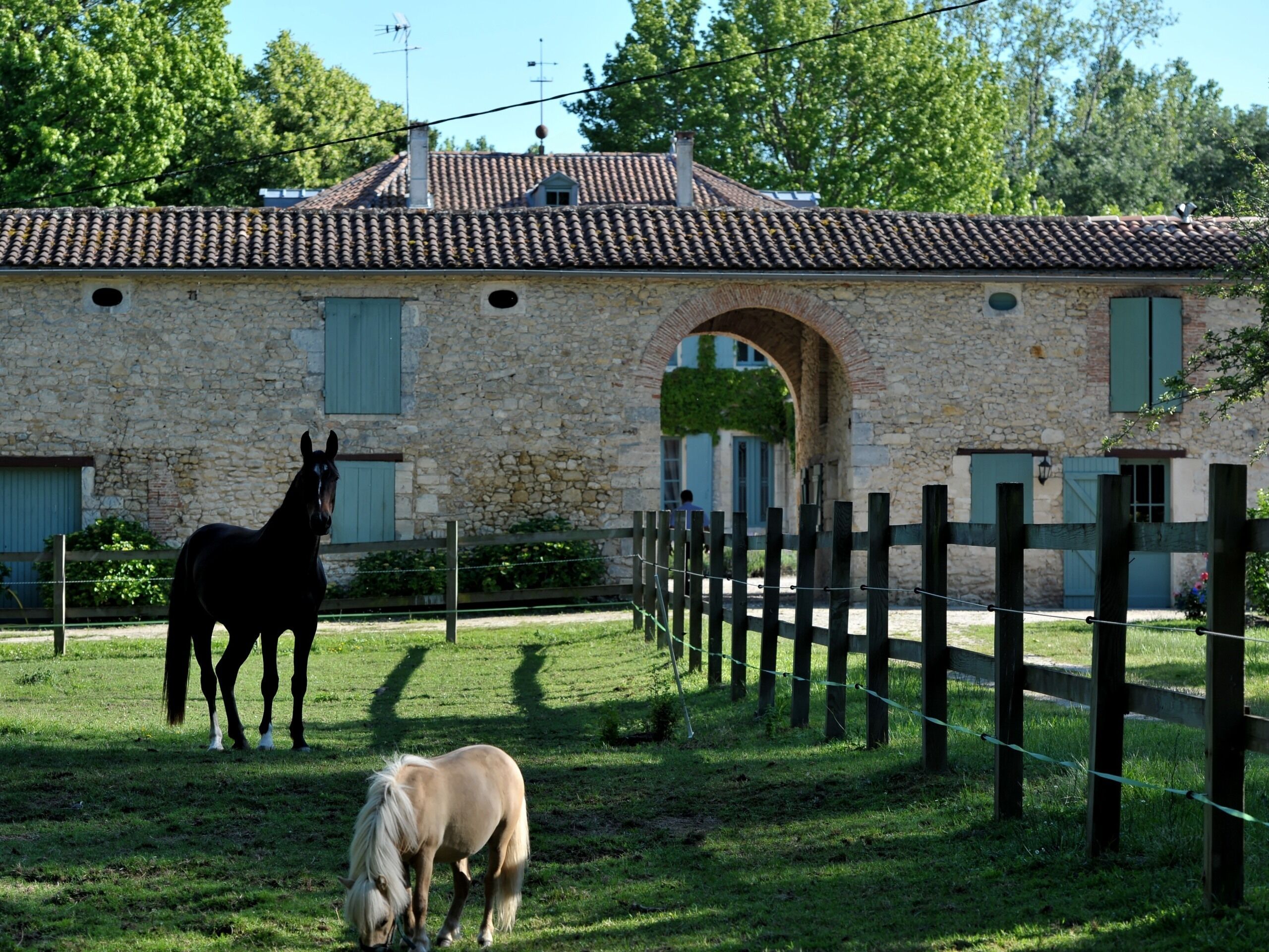 A stable with two horses, a stone building with a tiled roof, and a wooden fence.